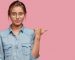 Waist up shot of pleasant looking woman in eyewear, points with thumb at free space, models against pink background, dressed in denim shirt. European young female demonstrates something aside